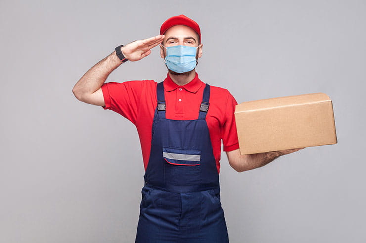 Retour au travail après la COVID-19 : un jeune homme portant un masque chirurgical, un uniforme bleu et un polo rouge se tient debout devant un fond gris, une boîte de carton à la main