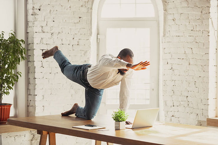 A business man doing a yoga pose on his desk in front of his laptop.