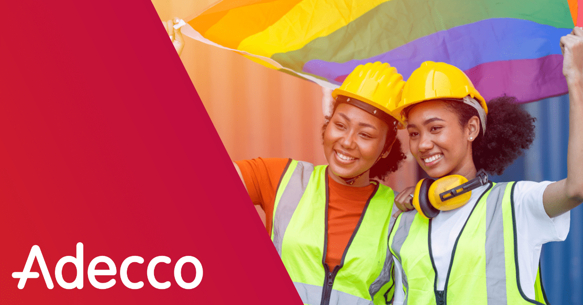 Two construction workers in hard hats and safety vests stand next to one another smiling. They are holding a rainbow flag over their heads