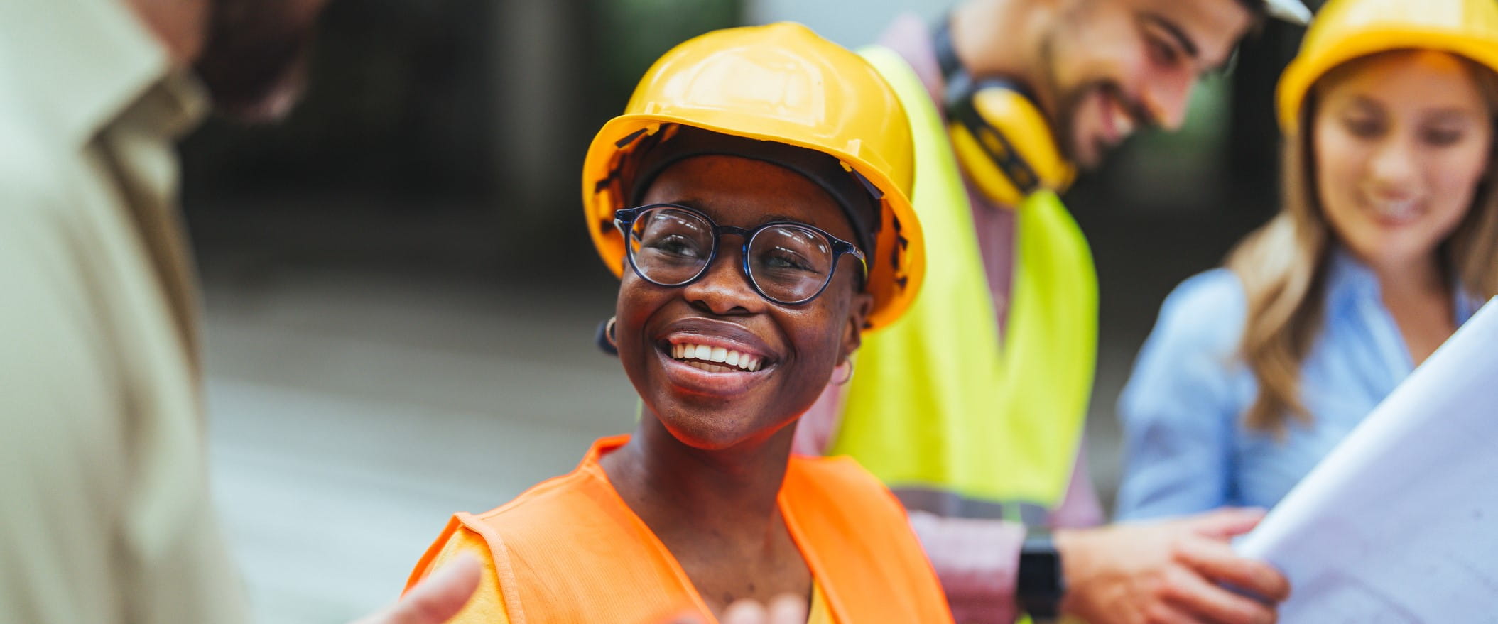 Worker in a hardhat and safety vest stands in a crowd of similarly dressed workers, smiling.