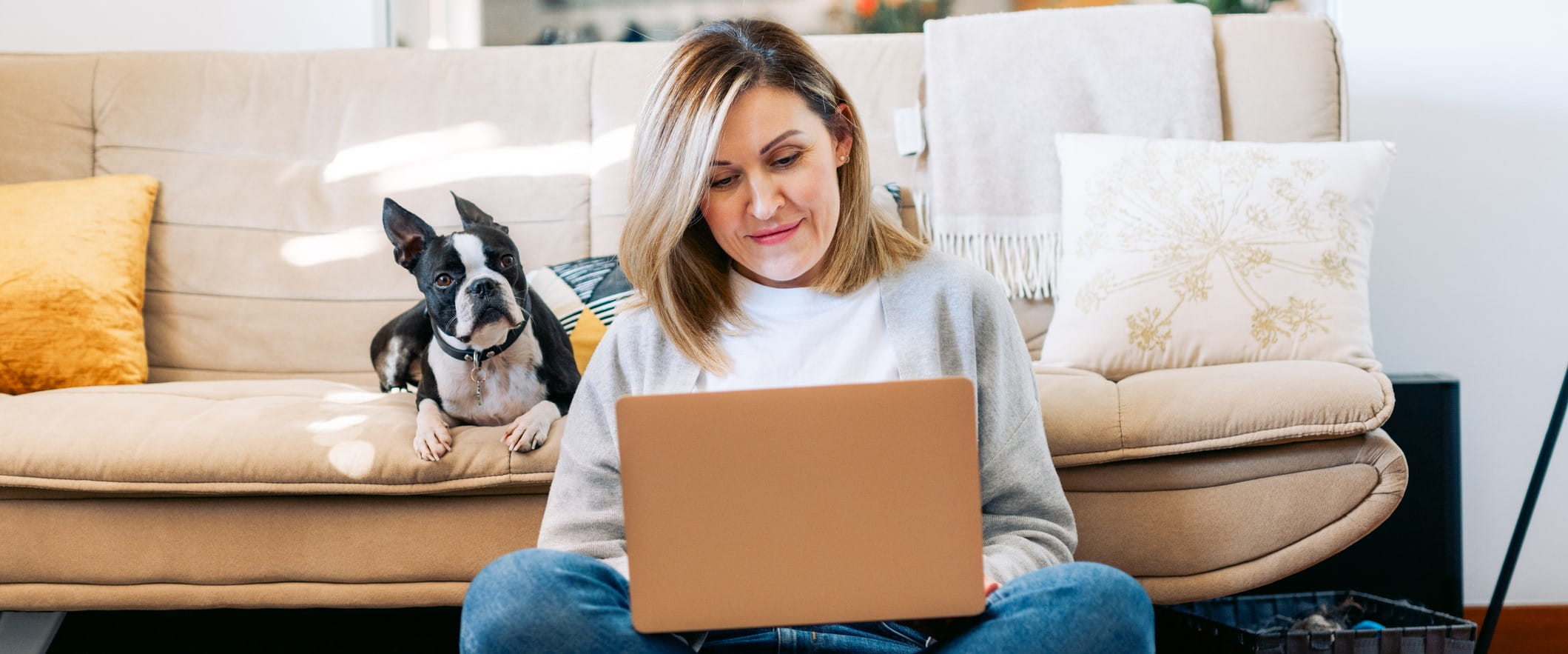 A person sits on the floor, leaning against a couch, with their laptop in their lap. A dog sits behind them on the couch.